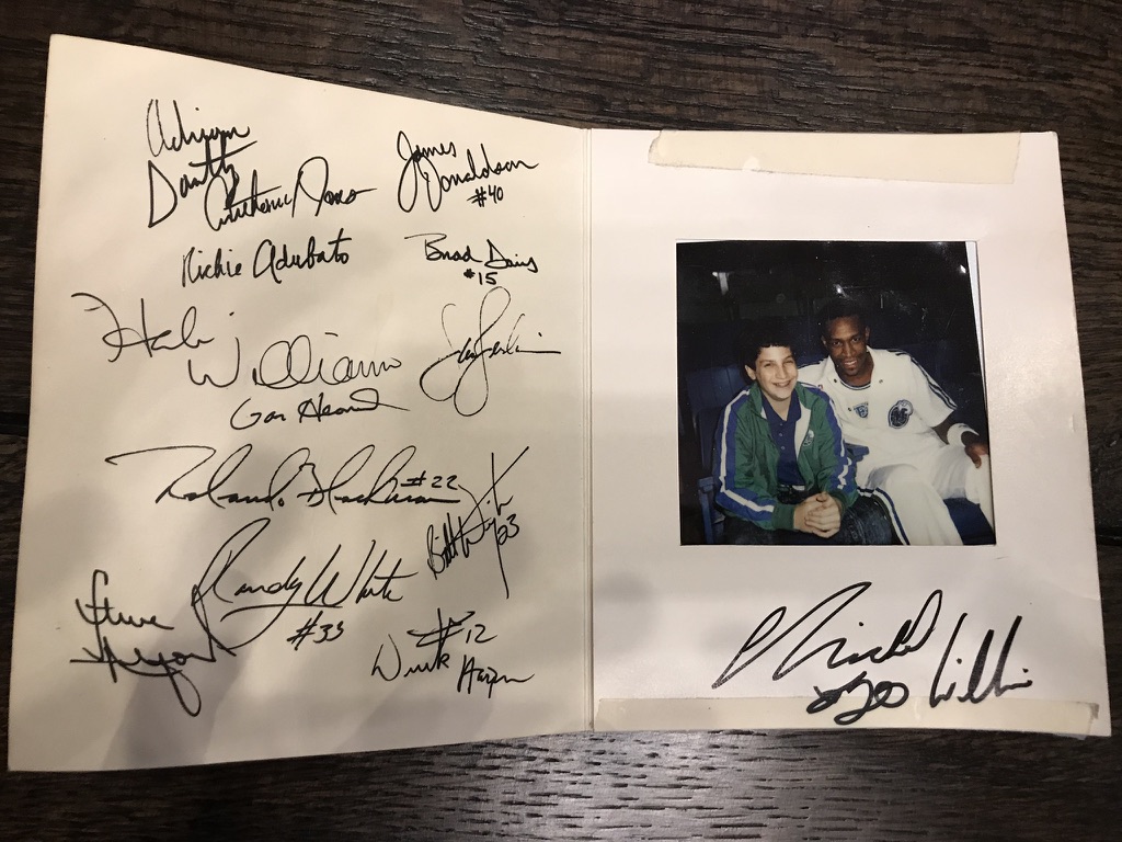 Signed Dallas Mavericks card from Andrew Hillman's childhood as a promotions assistant and ball boy. Childhood photo of Andrew with a Mavericks player; Herb Williams autograph under the photo, plus other team signatures.