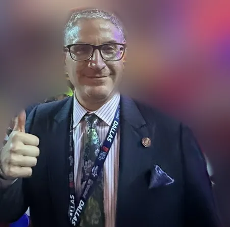 Andrew Hillman in a navy suit and Dallas lanyard giving a thumbs-up on a convention floor