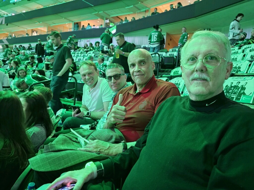 Andrew Hillman with Sergei Gonchar and Maj. Gen. James L. Williams (Ret.) in arena seats at a Dallas Stars game. Green arena lighting. A Dallas Stars jersey, PAVELSKI 16, is visible in the background. Seat signs say PLAYOFF HOCKEY and ROUND 2 GAME 5.