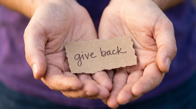 Open hands holding a torn paper note reading give back, illustrating Andrew Hillman's giving